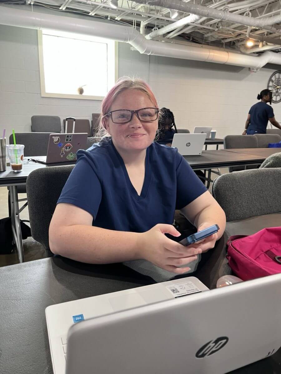 A student in navy scrubs smiling at her laptop during a CNA class